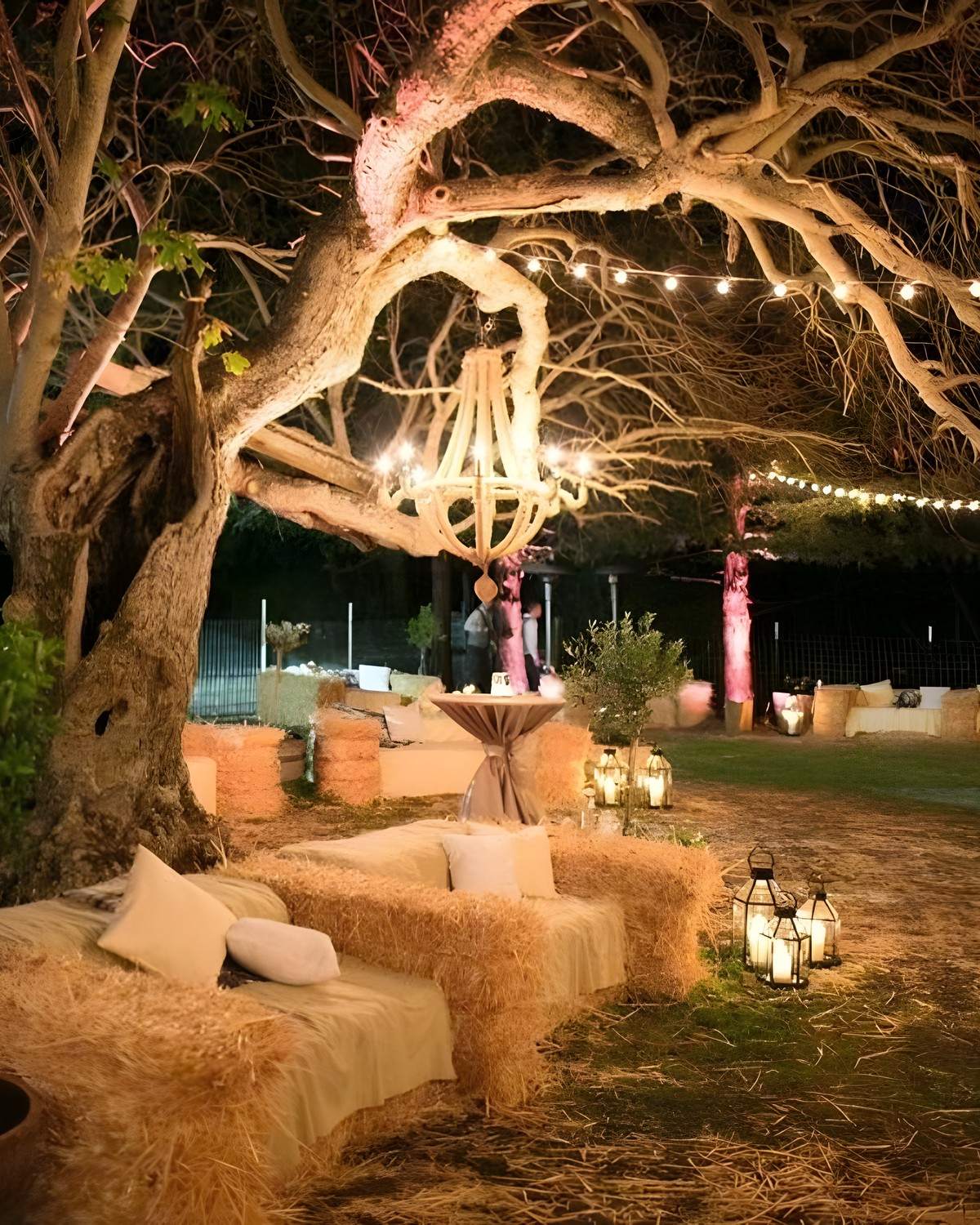 A straw-bale lounge under a lit olive tree, with a chandelier and lanterns, in the evening.